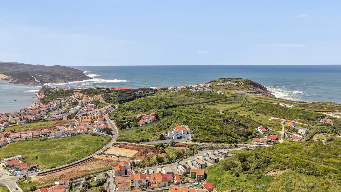 Aerial view of a coastal town with red-roofed houses, green hills, and blue ocean under a clear sky.