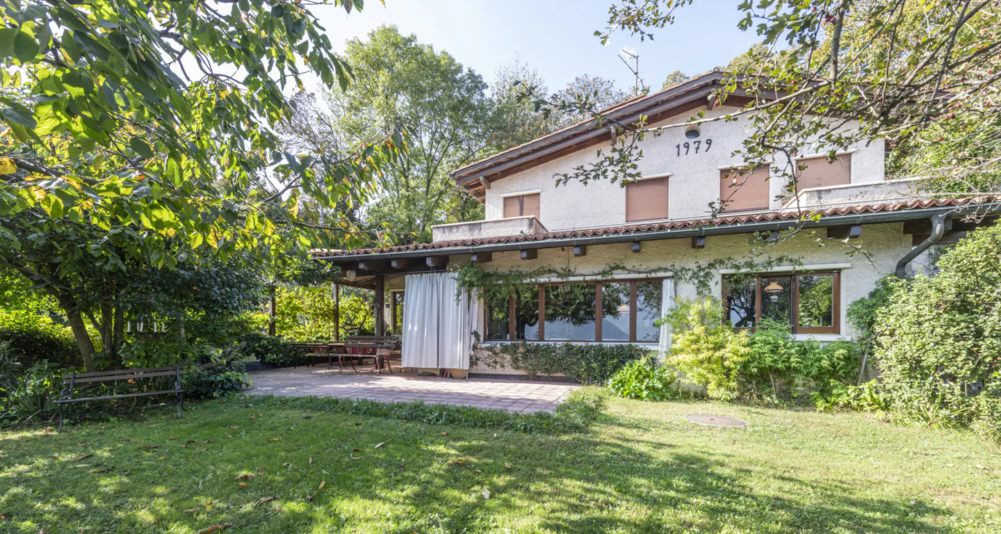 Exterior view of a two-story house with a lawn, patio, and trees. The house is light beige with brown shutters and a brown tile roof. The year "1979" is above the second-story windows.