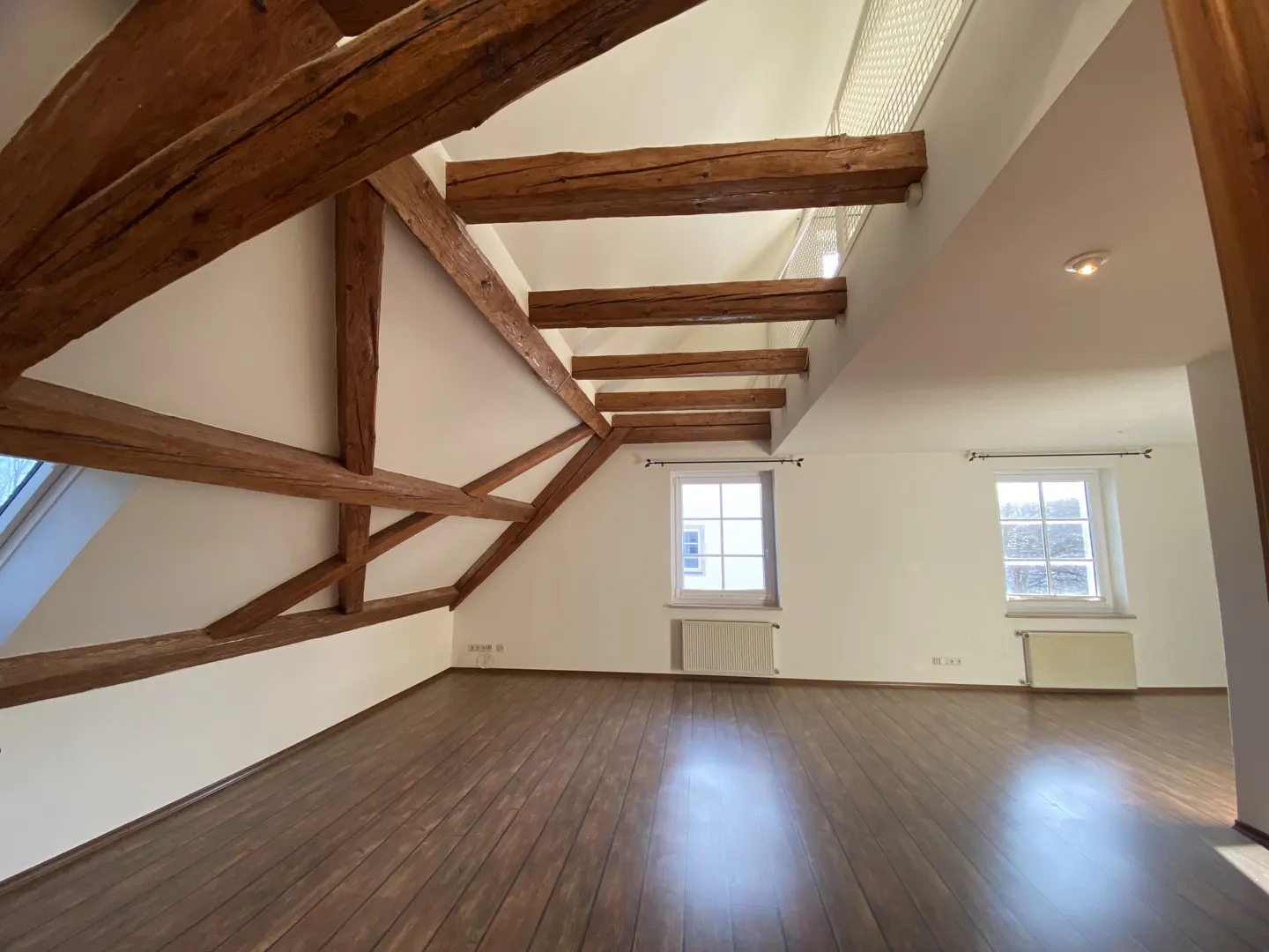 Attic room with dark wood flooring, white walls, exposed wooden ceiling beams, and two windows with radiators below.