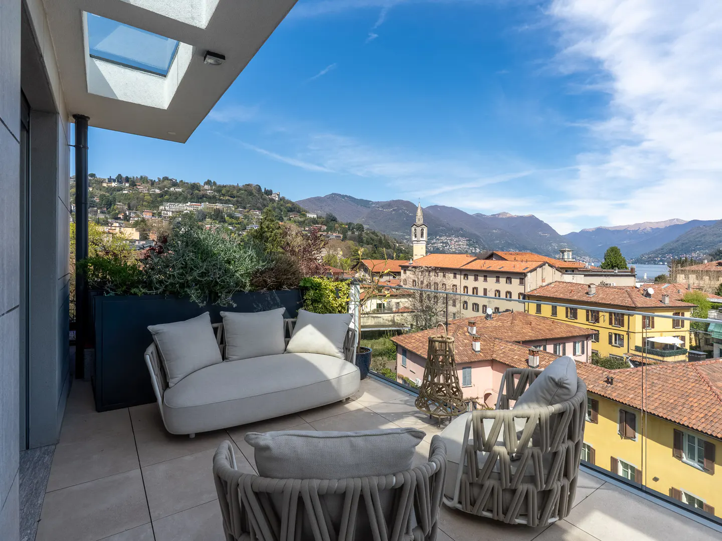 Balcony with a white sofa and chairs overlooks a European town with orange rooftops, mountains, and a blue sky.