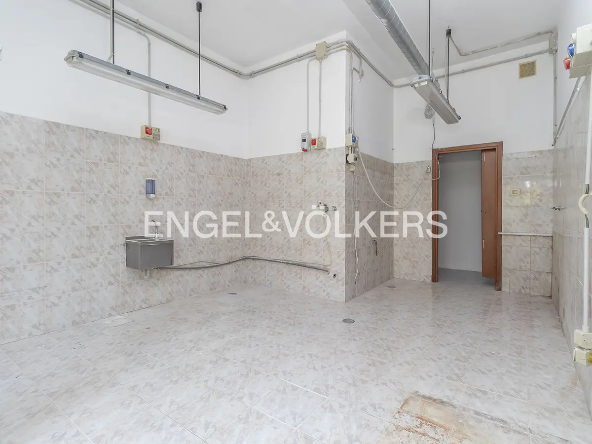 Empty commercial space with tiled walls and floor, sink, fluorescent lights, and a doorway. The Engel & Völkers logo is superimposed.