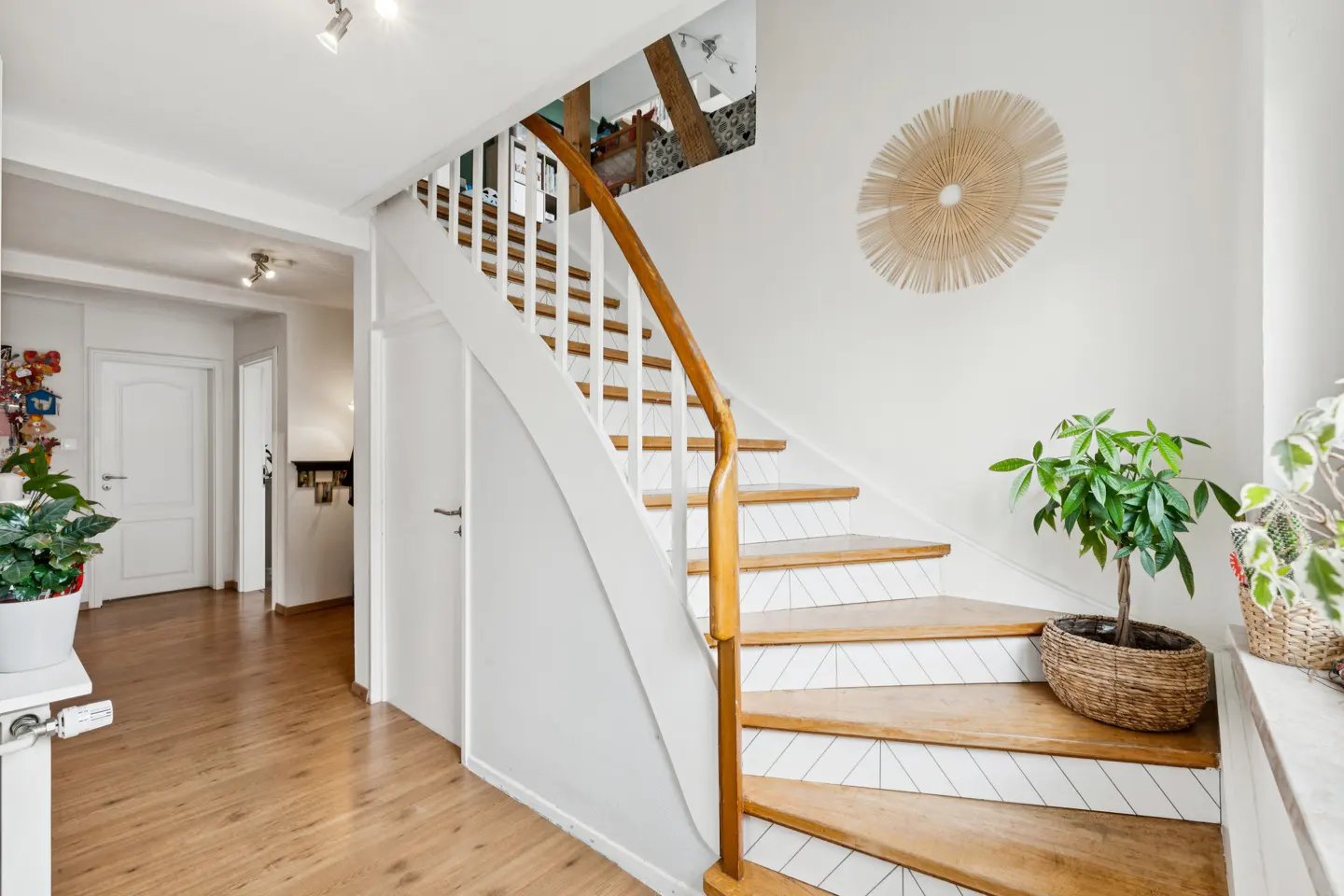 Bright hallway with wooden floors, white walls, and a staircase with a wooden handrail. A round straw decoration hangs on the wall.