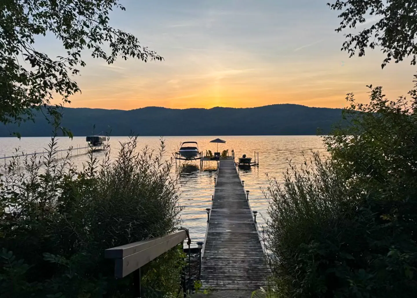 Sunset view of a lake with a wooden dock leading to boats and chairs, framed by trees.
