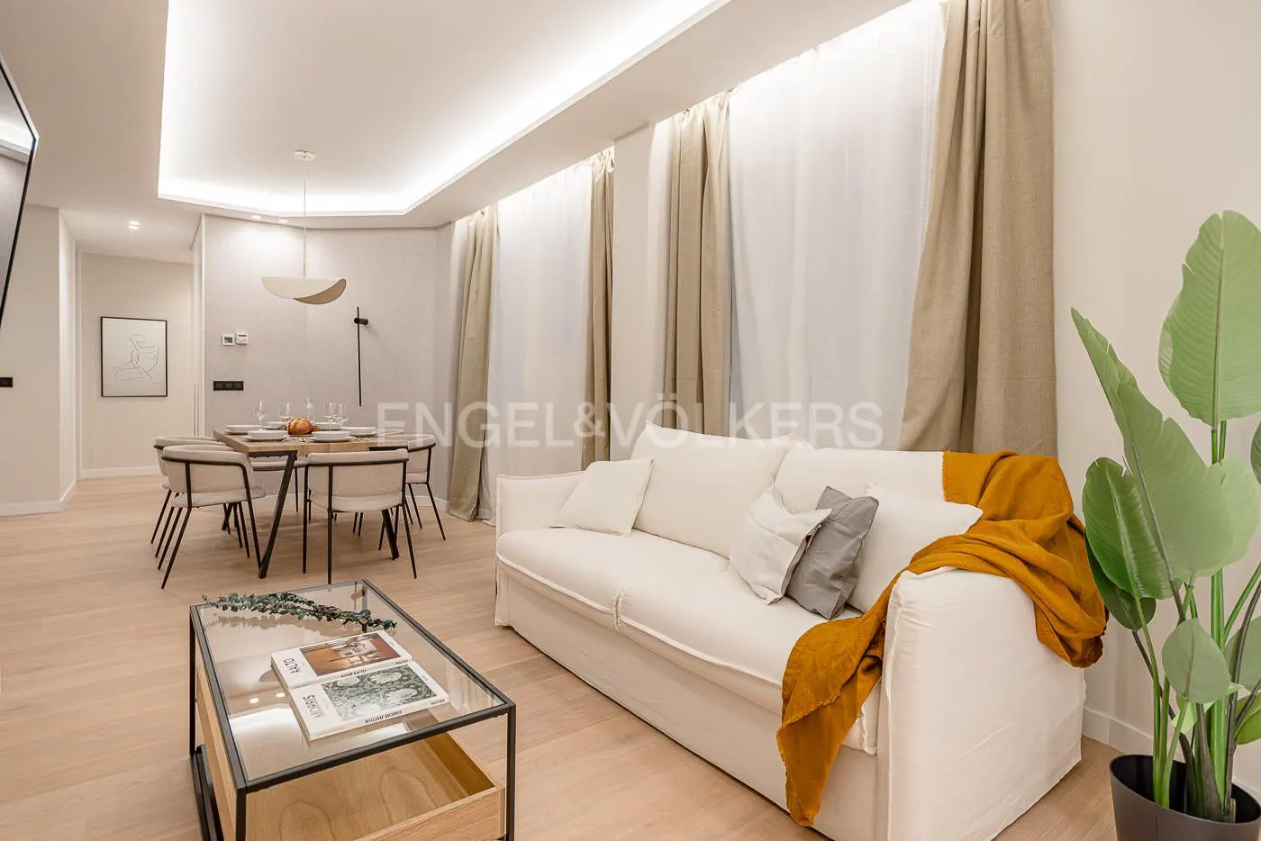 Bright living room with a white sofa, tan curtains, and a dining table set for a meal. A glass coffee table sits in front of the sofa.