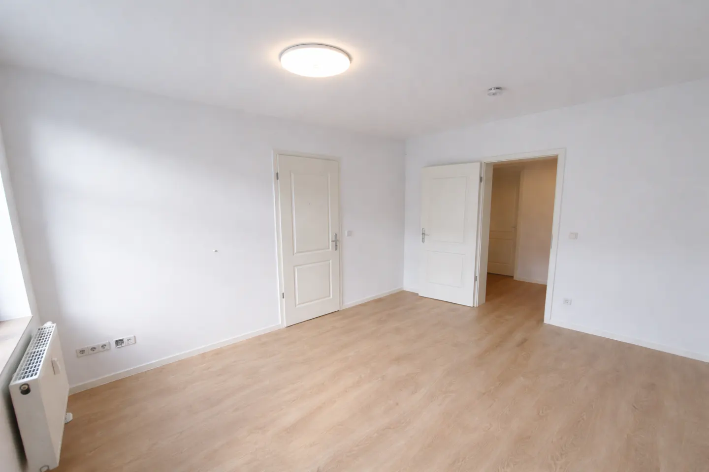 Bright, empty room with light wood floors, white walls, and two white doors. A radiator sits by the window.