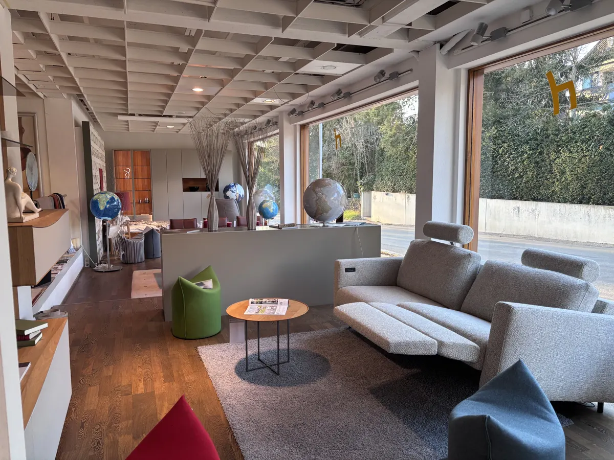 Showroom interior with a gray sofa, round table, and globes. Large windows offer a view of greenery. Modern design with a grid ceiling.