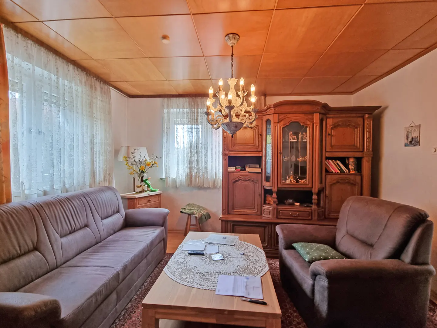 Living room with brown sofa, armchair, wood cabinet, and coffee table with lace doily. Chandelier hangs from wood paneled ceiling.