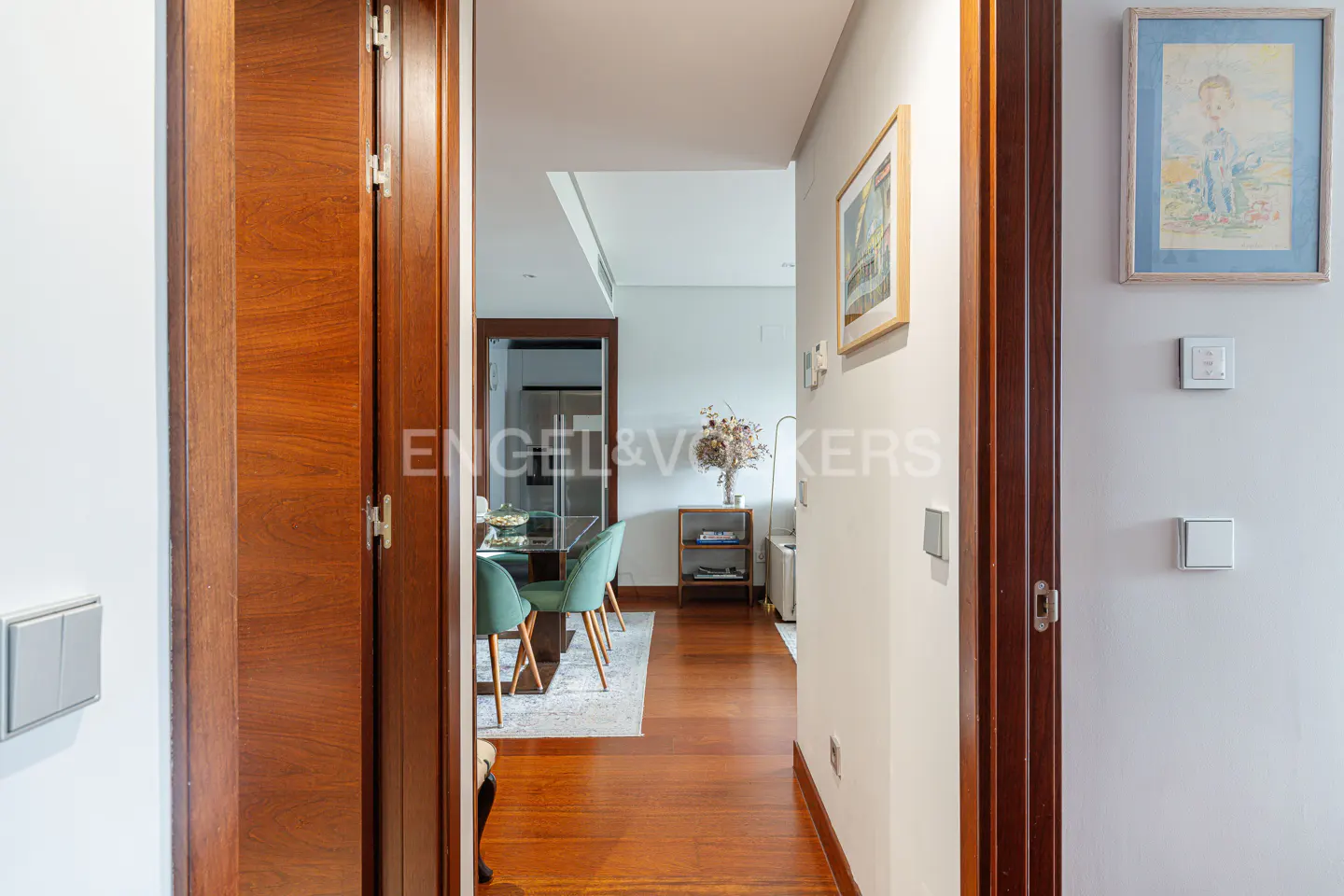 A view through a doorway into a modern hallway with hardwood floors and a dining area with green chairs.