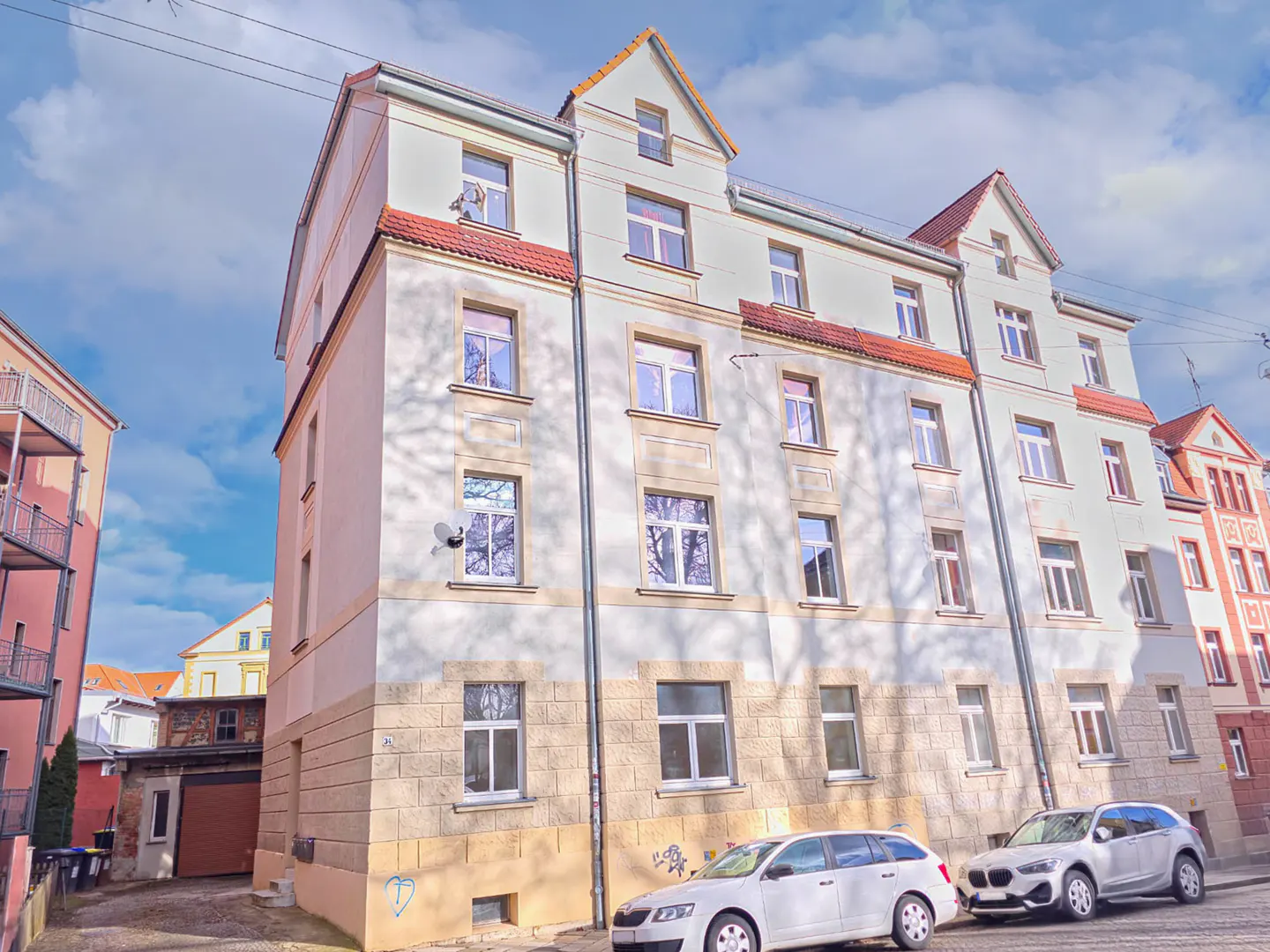 Exterior view of a four-story apartment building with white and tan facade, red roof, and two cars parked in front.