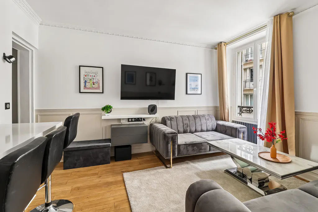 Modern living room with a grey velvet sofa, marble coffee table, wall-mounted TV, and large window with tan curtains.