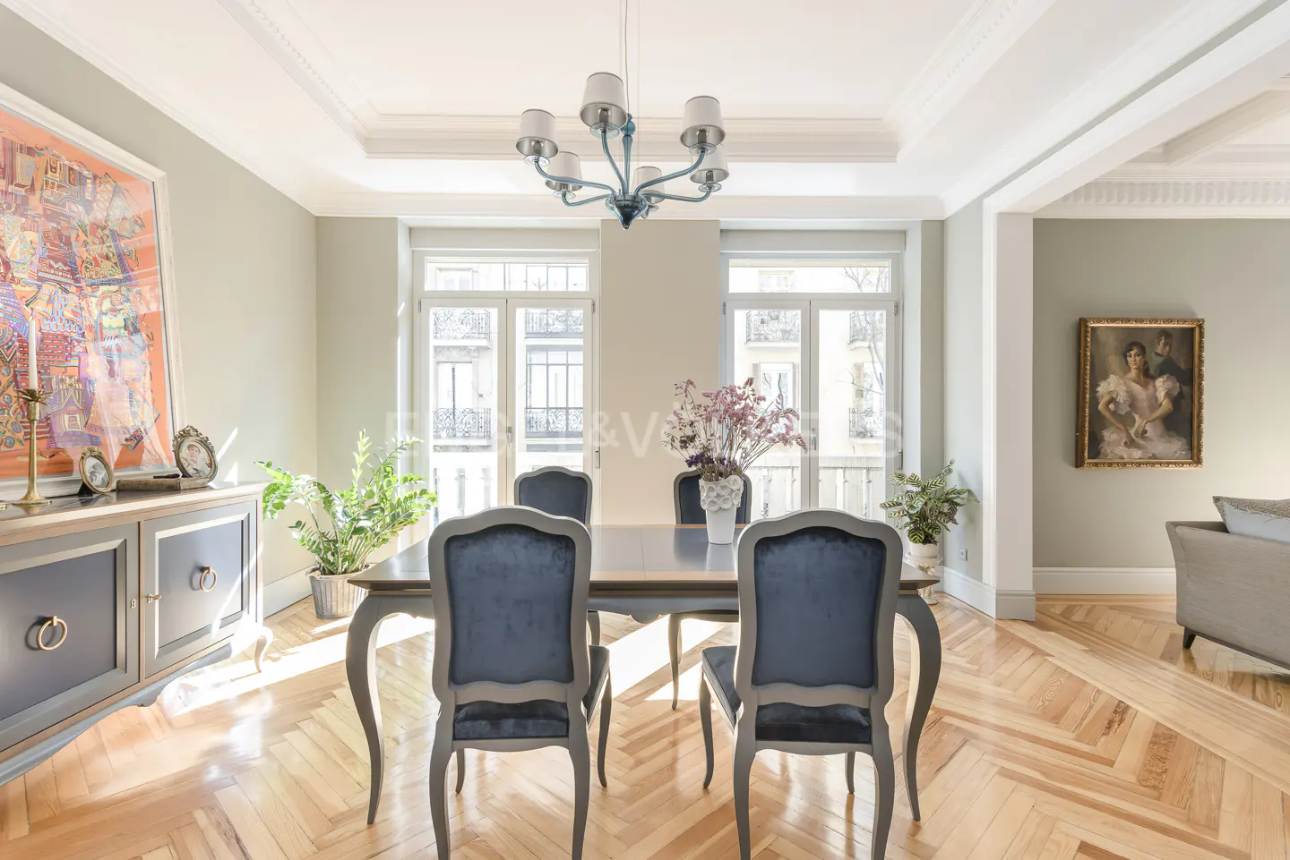 Bright dining room with parquet floors, blue chairs around a table, and a chandelier. Large windows let in natural light.