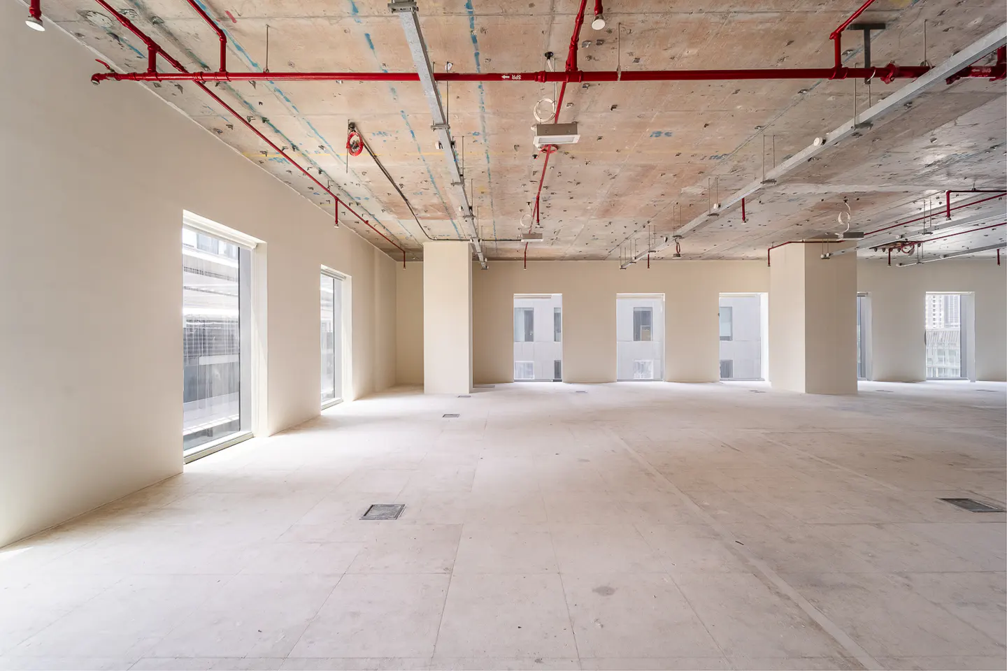 Unfinished office space with concrete floors and ceiling, red pipes, and large windows.