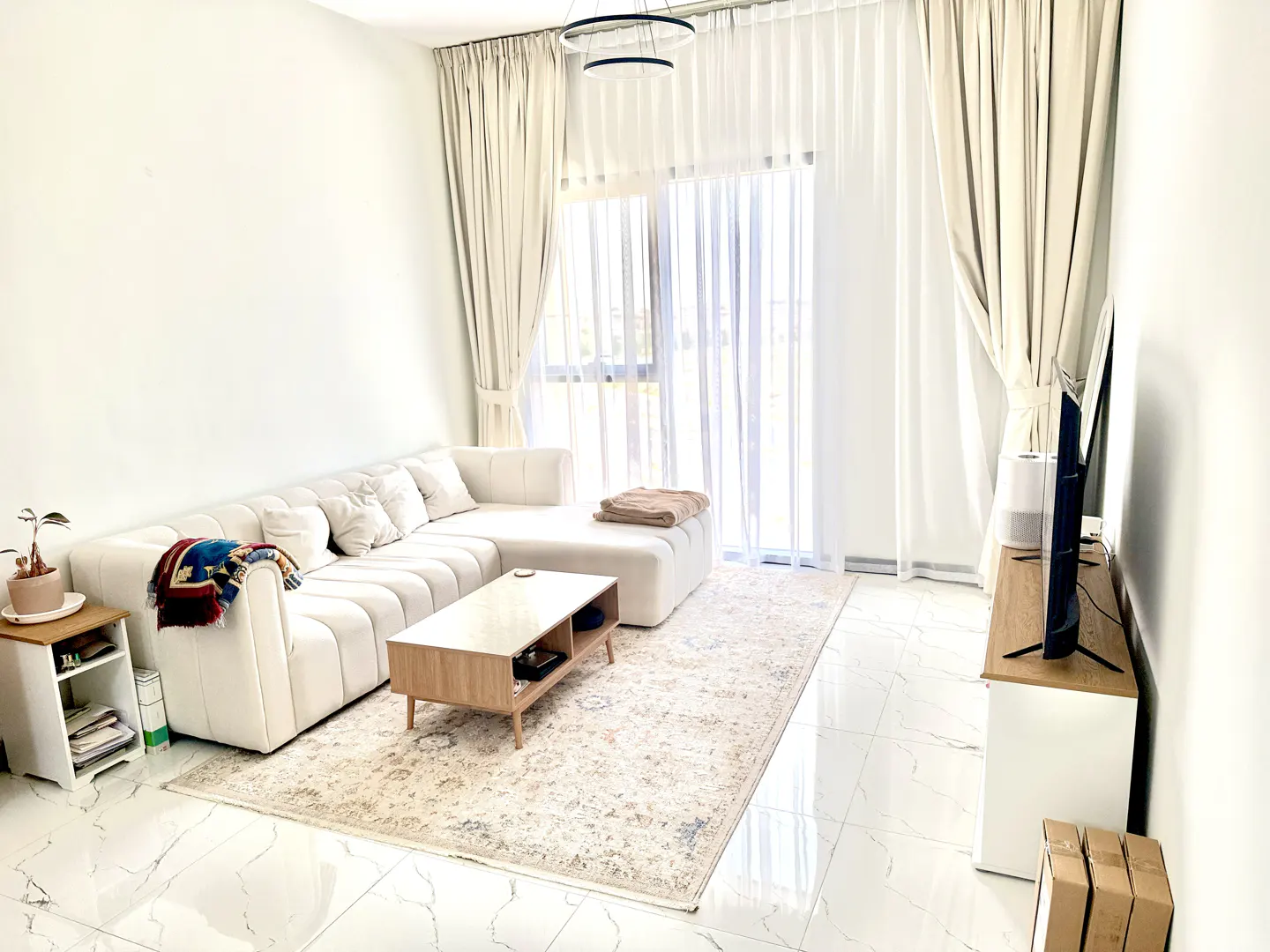Bright living room with white walls, marble floors, and a cream sectional sofa. A light wood coffee table sits on a patterned rug. Sheer curtains adorn a window.