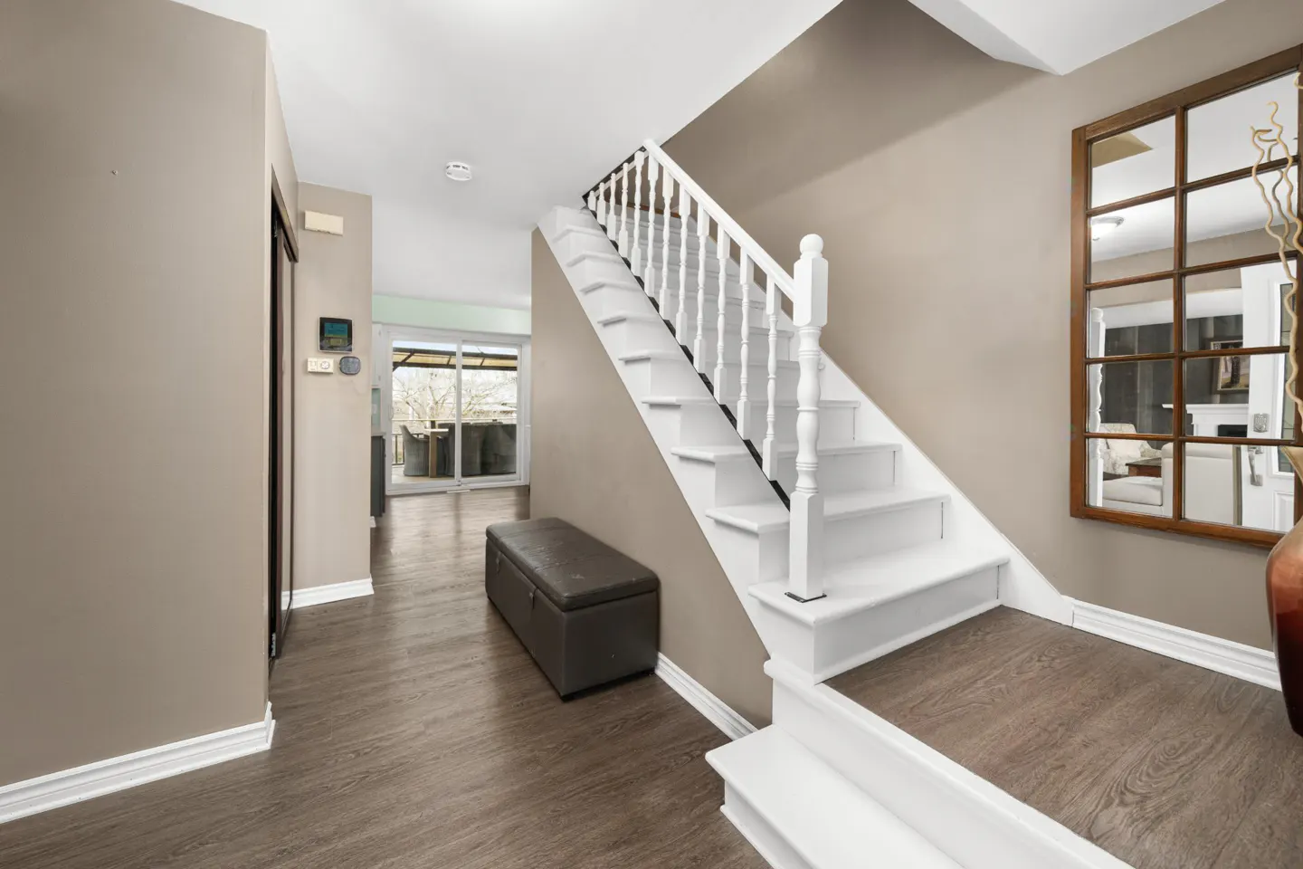 Entryway with white stairs, a brown storage bench, and a large grid mirror on a beige wall. Wood-look flooring and a glimpse of the backyard.