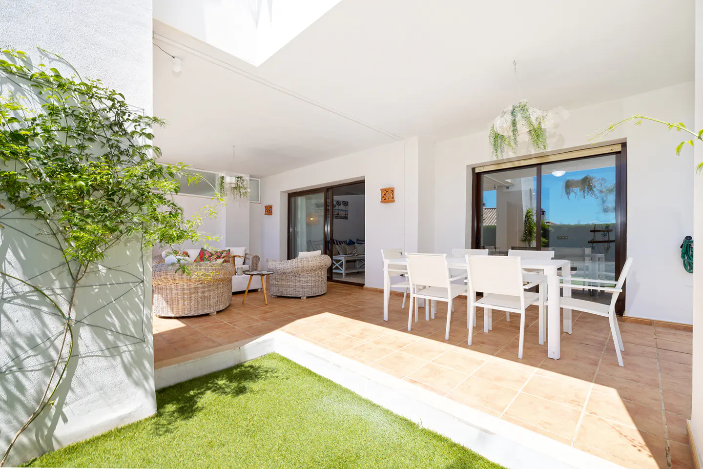A bright patio with white walls, tile flooring, and a green lawn area. Wicker furniture and a white dining set are visible.