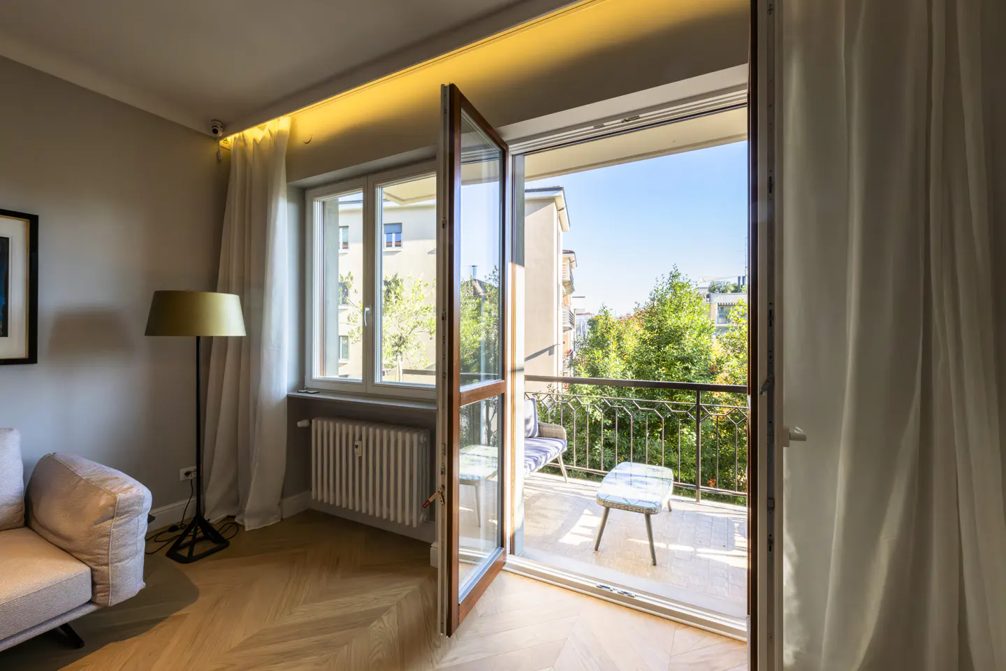 Living room with open glass doors to a balcony. Balcony has a chair and table. Trees and buildings are visible in the background.
