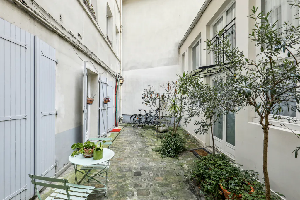 A narrow courtyard with stone pavers, white walls, and blue shutters. A small table and chairs sit near a door, and a bicycle is parked further back.