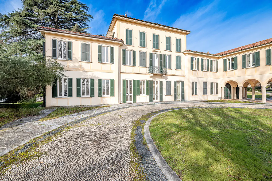 Exterior view of a large, light yellow house with green shutters and a cobblestone driveway.