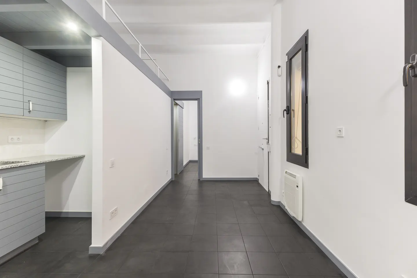 A bright, modern apartment interior with gray cabinets, white walls, and dark tile floors. A hallway leads to another room.