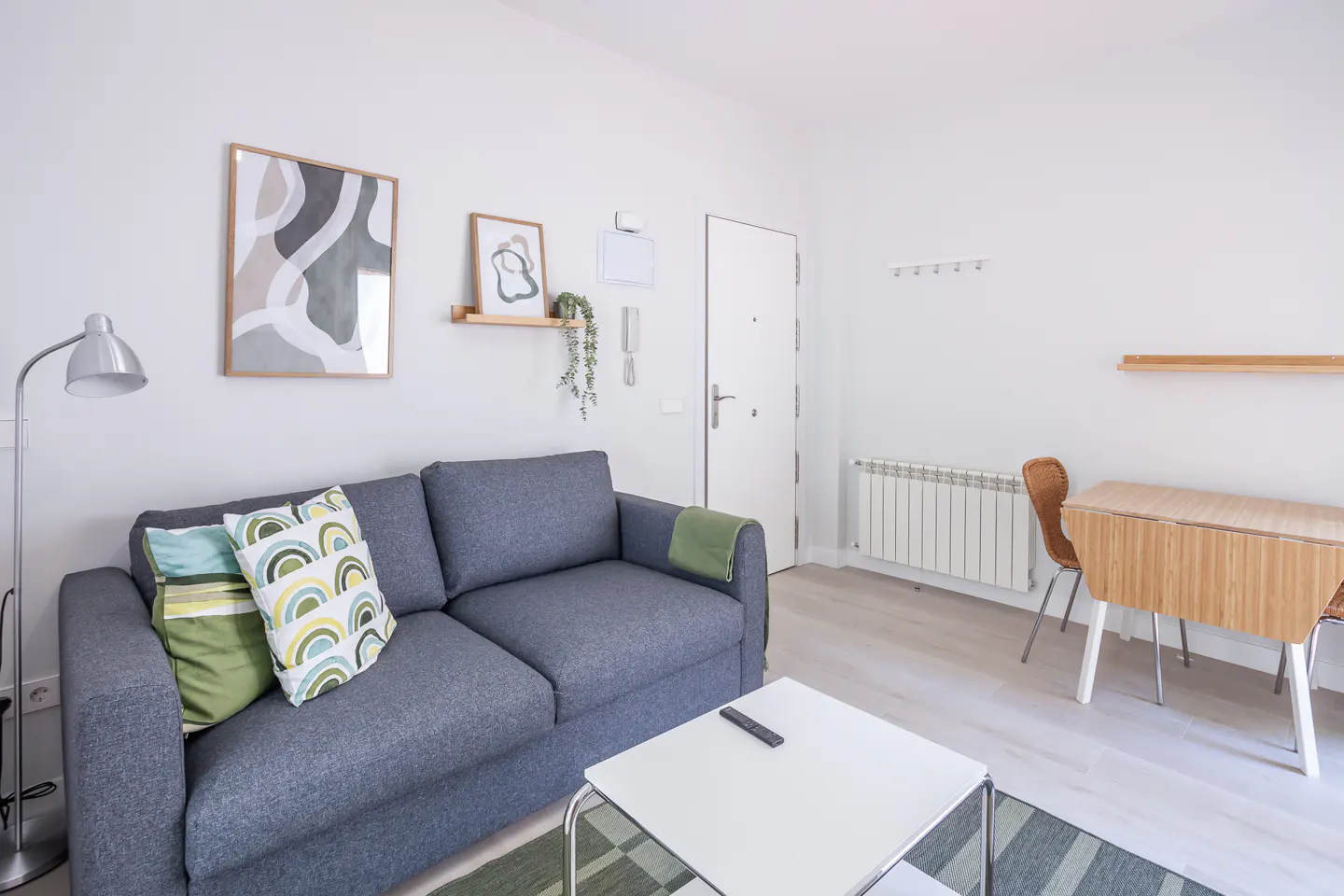 Bright living room with a gray sofa, colorful pillows, a white coffee table, and a wooden dining table with chairs. Artwork adorns the white walls.