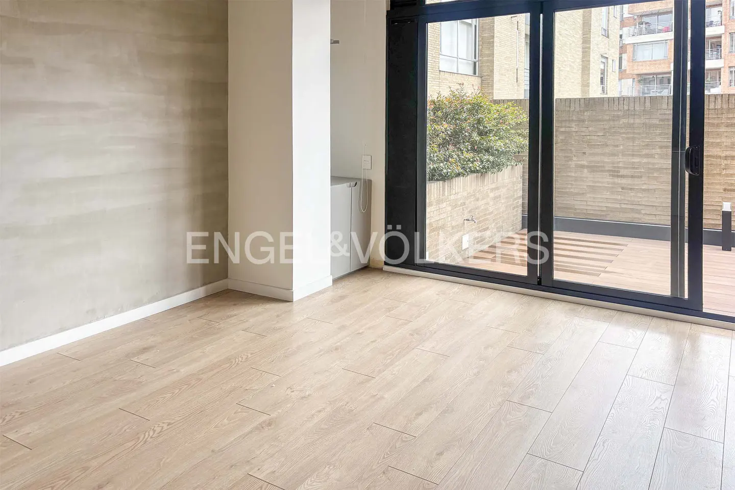An empty room with light wood floors, a textured beige wall, and a black-framed sliding glass door to a balcony.