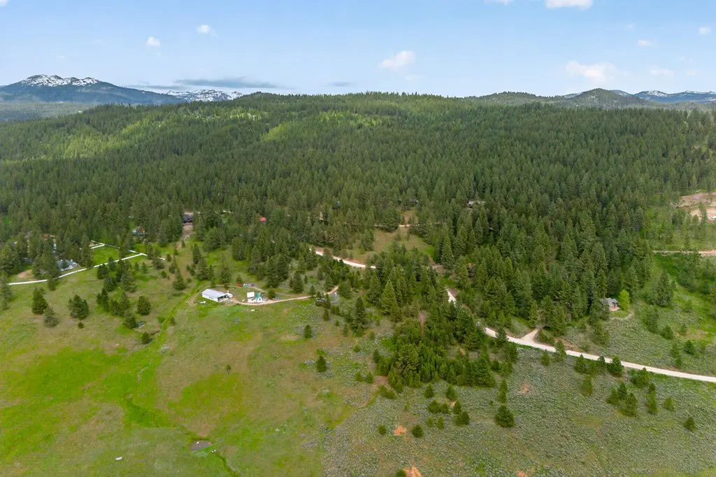 Aerial view of a green, tree-covered landscape with a dirt road, buildings, and snow-capped mountains in the distance under a blue sky.