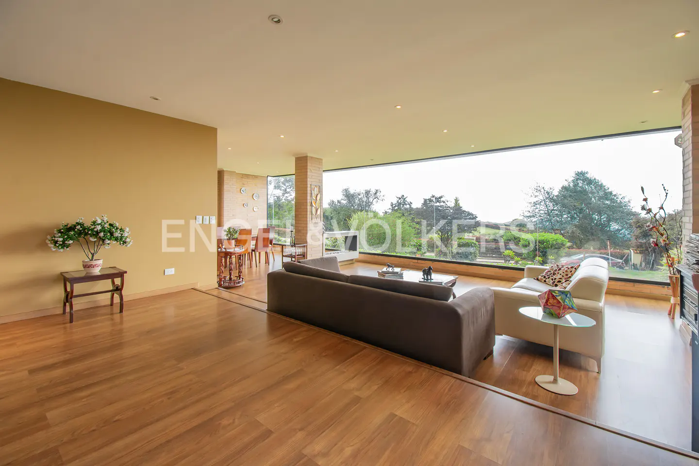 A bright living room with wood floors, a brown sofa, and a white chair facing a large window with a view of trees.