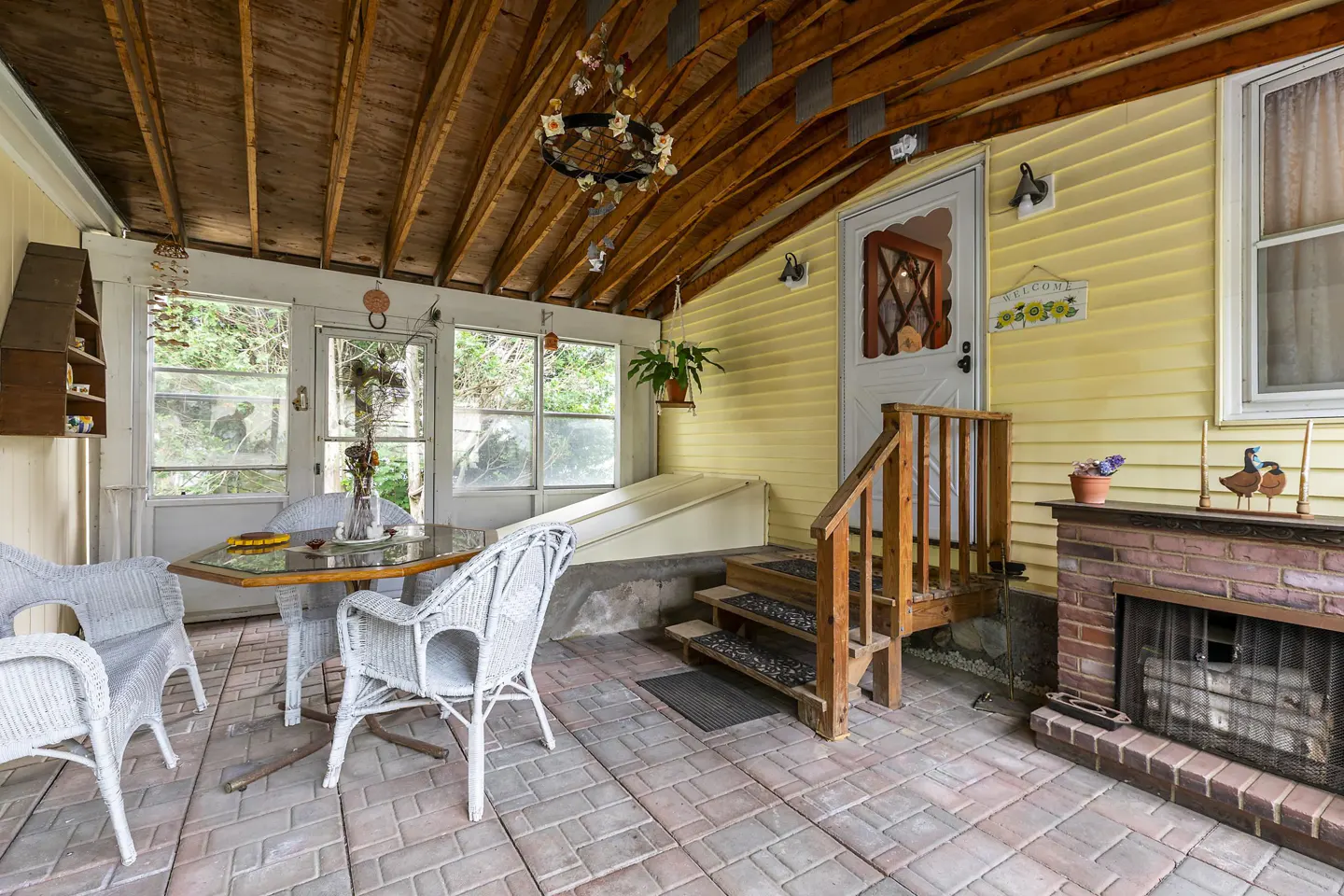 Enclosed porch with brick floor, yellow siding, and exposed wood ceiling. Wicker furniture, brick fireplace, and steps to a white door.