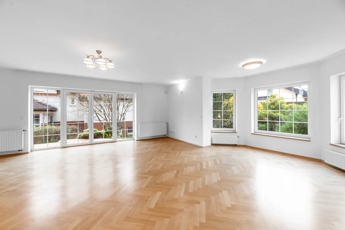 Bright, empty room with herringbone wood floors, white walls, and large windows showing trees and houses outside. A chandelier hangs from the ceiling.