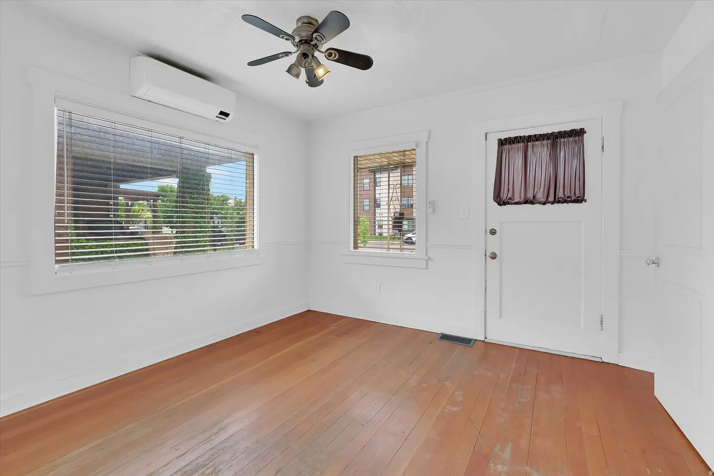 Bright room with white walls, hardwood floors, a ceiling fan, wall-mounted AC unit, two windows, and a door with a curtain.