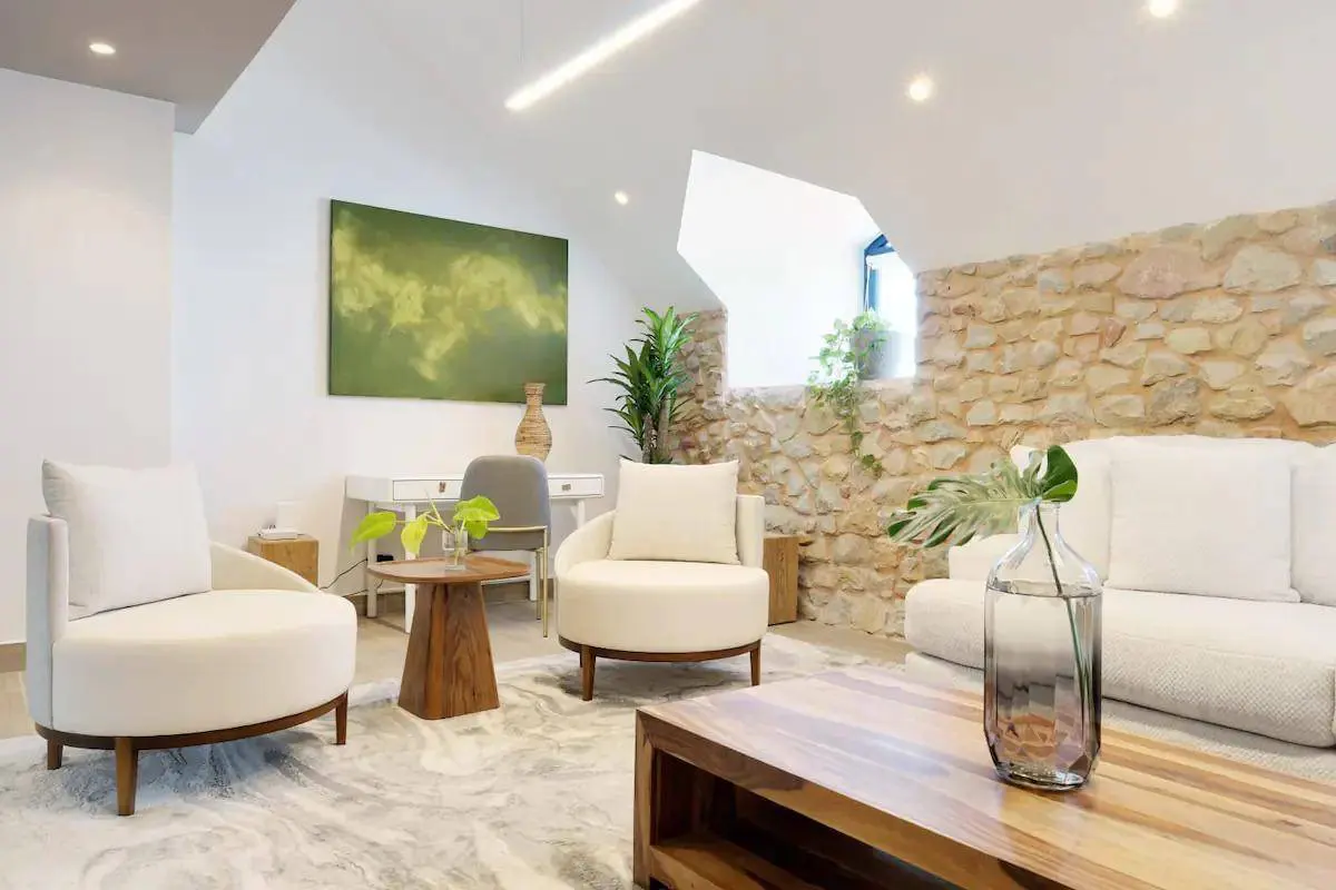Living room with white armchairs, sofa, and stone wall. A wooden coffee table and desk add warmth to the bright, modern space.