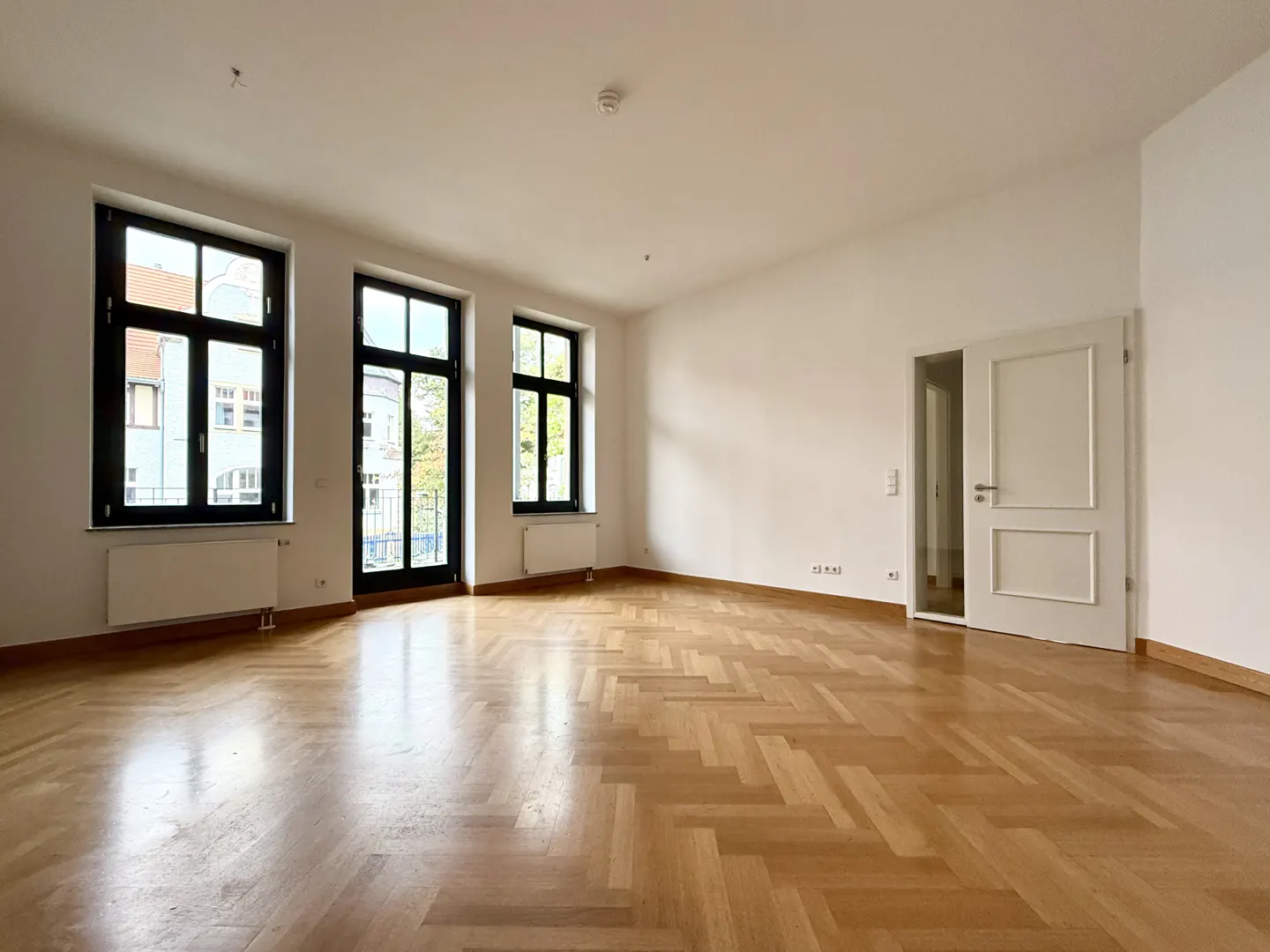 Bright, empty room with herringbone wood floors, white walls, and black-framed windows. A white door is open to another room.