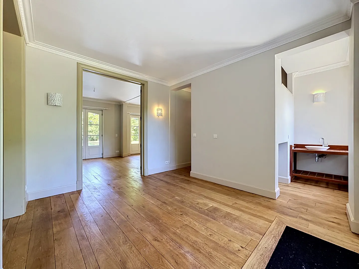 Interior view of a home with hardwood floors, light gray walls, white trim, and doorways leading to other rooms. A sink and vanity are visible in one room.