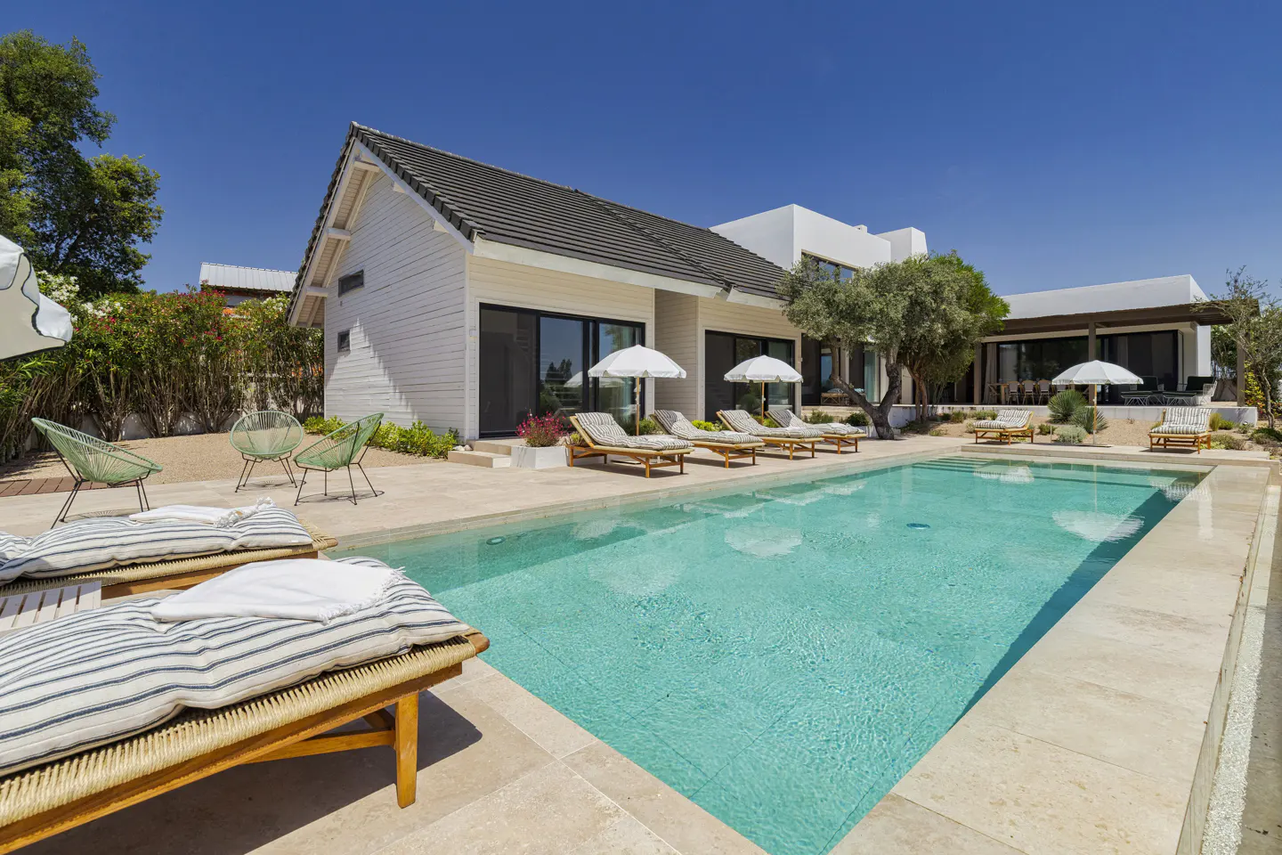 Modern white villa with a turquoise swimming pool, lounge chairs, and umbrellas on a sunny day.