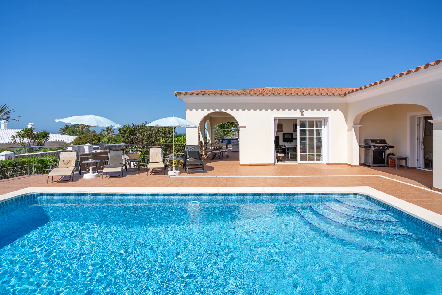 A bright blue swimming pool in front of a white house with lounge chairs and umbrellas.