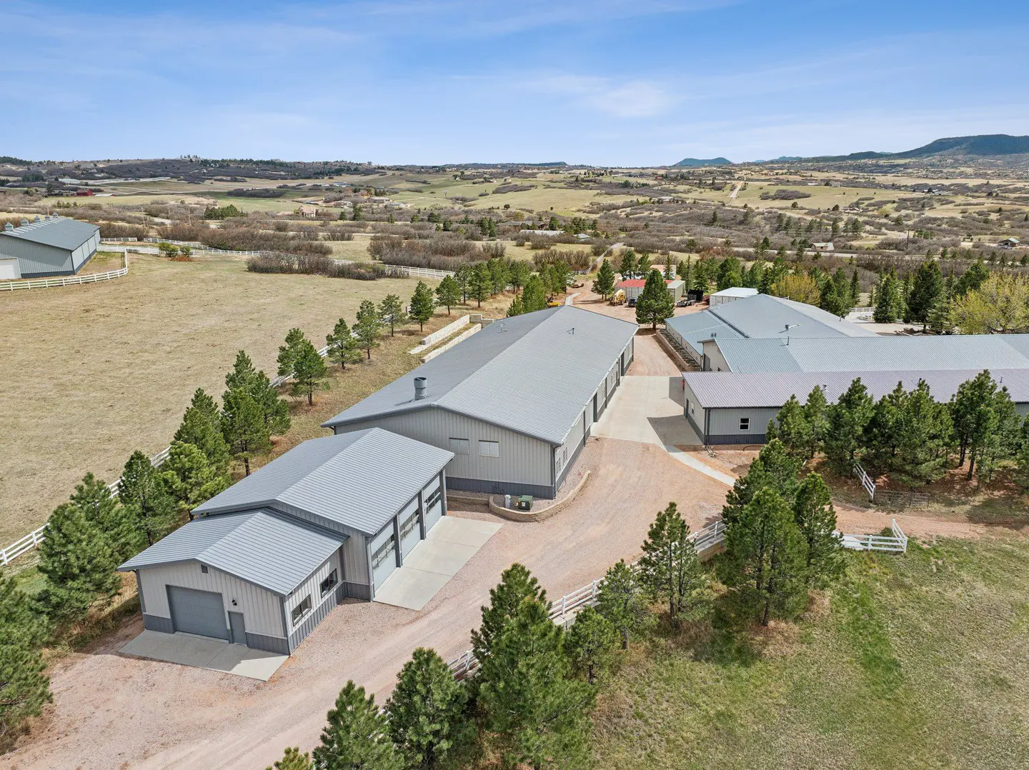 Aerial view of a large property with multiple gray metal buildings, green trees, and a vast landscape under a blue sky.