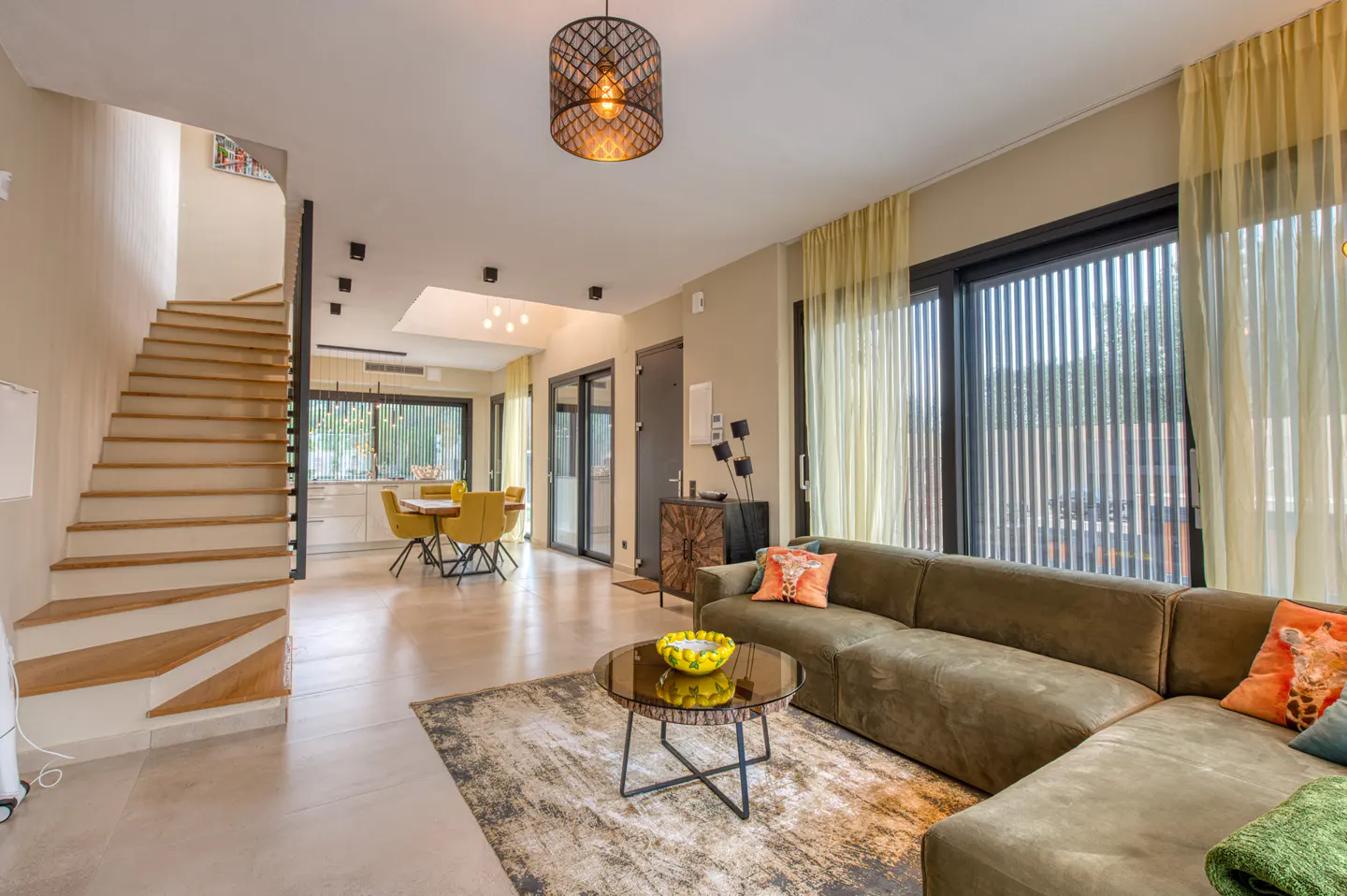 A modern living room with a brown sectional sofa, round coffee table, and stairs. Yellow curtains and dining chairs add pops of color.