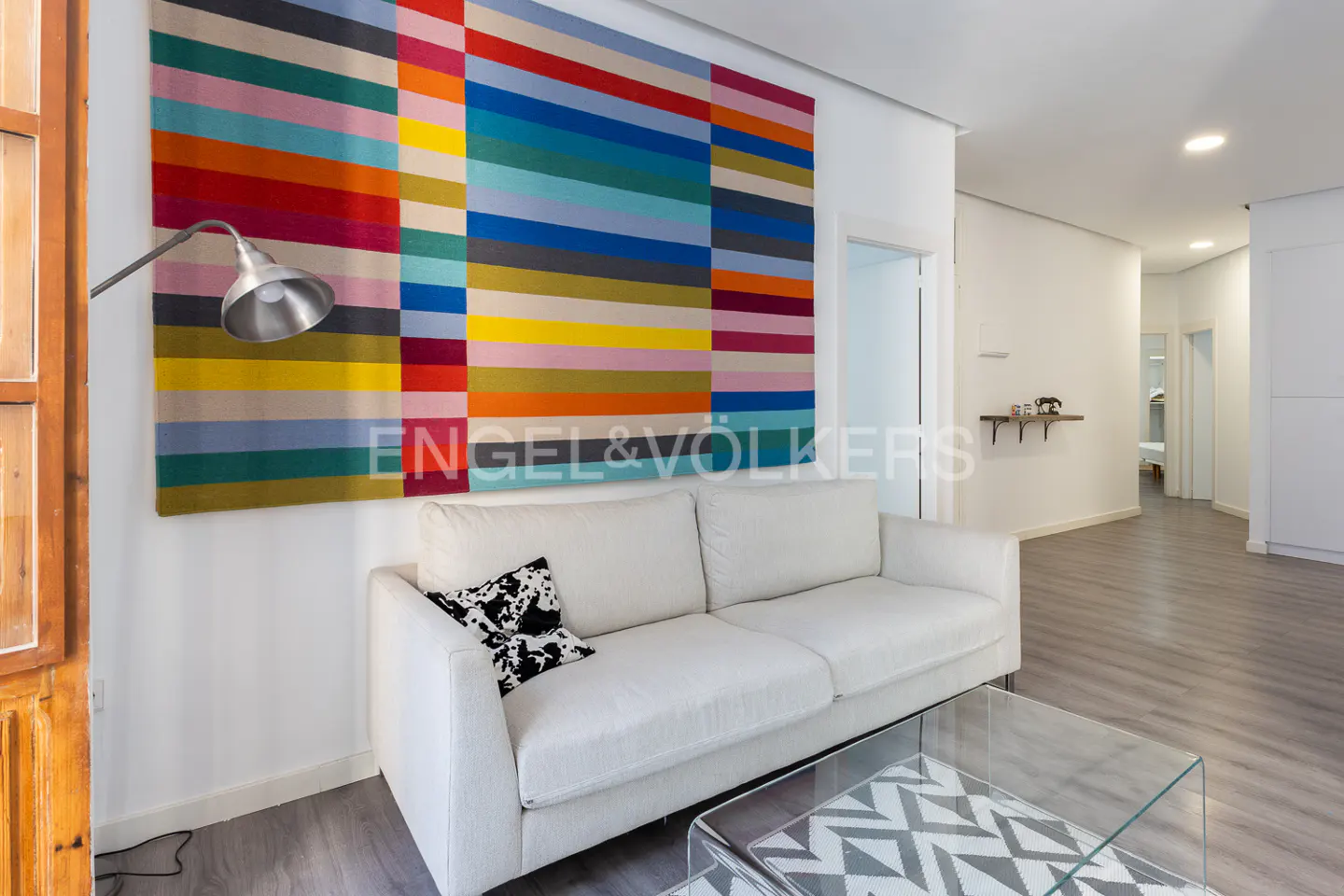 Living room with white sofa, glass table, and colorful striped wall art. Gray floors and a hallway are visible.