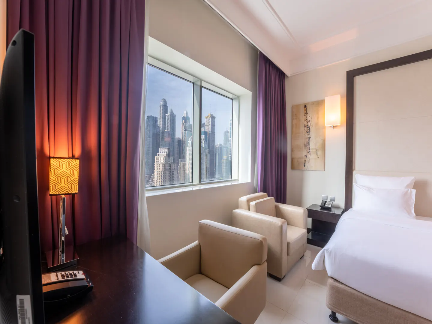 Hotel room with a bed, desk, and two chairs. A large window shows a city skyline. Purple curtains frame the window.