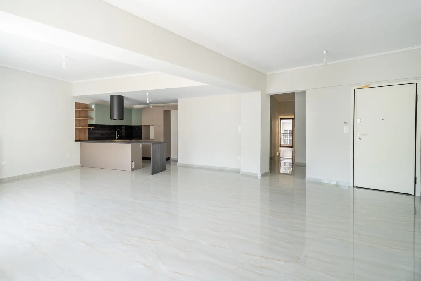 Bright, empty apartment with white walls and marble floors. An open kitchen with a black range hood is visible. A white door is on the right.
