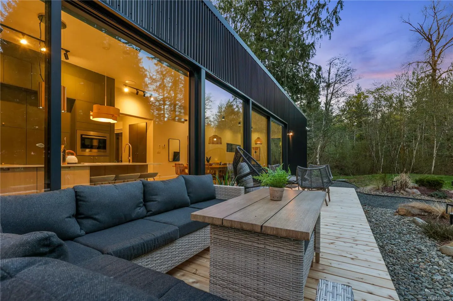 Modern black home with large windows, a wooden deck, and outdoor furniture. The interior features a kitchen and dining area. Trees in the background.