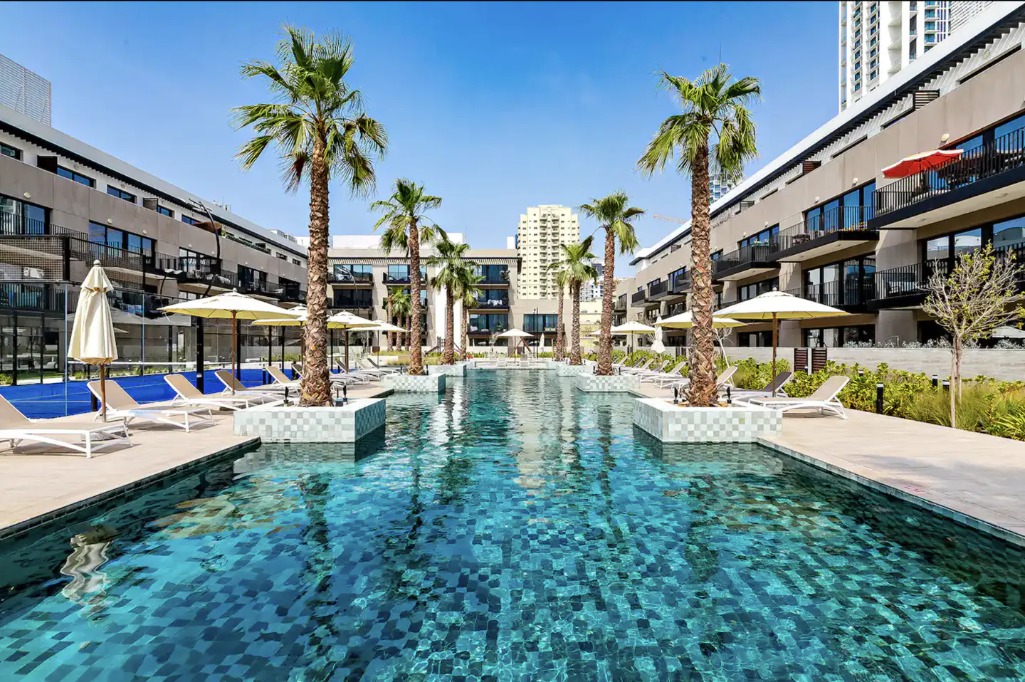 Outdoor pool with palm trees and lounge chairs between modern buildings under a blue sky.