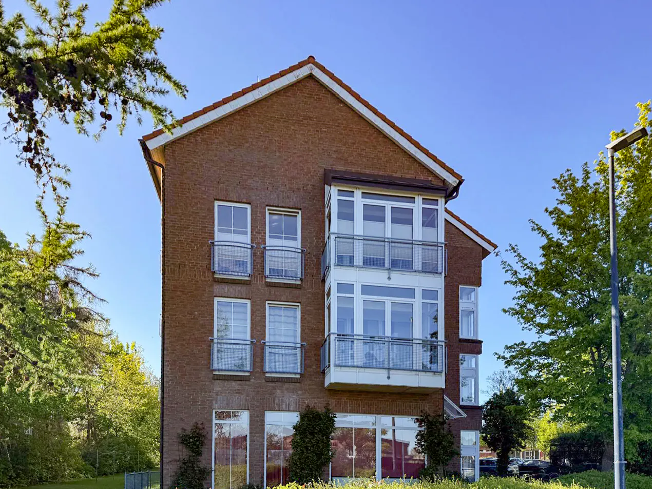 A three-story brick building with white-framed windows and glass balconies, surrounded by green trees under a blue sky.