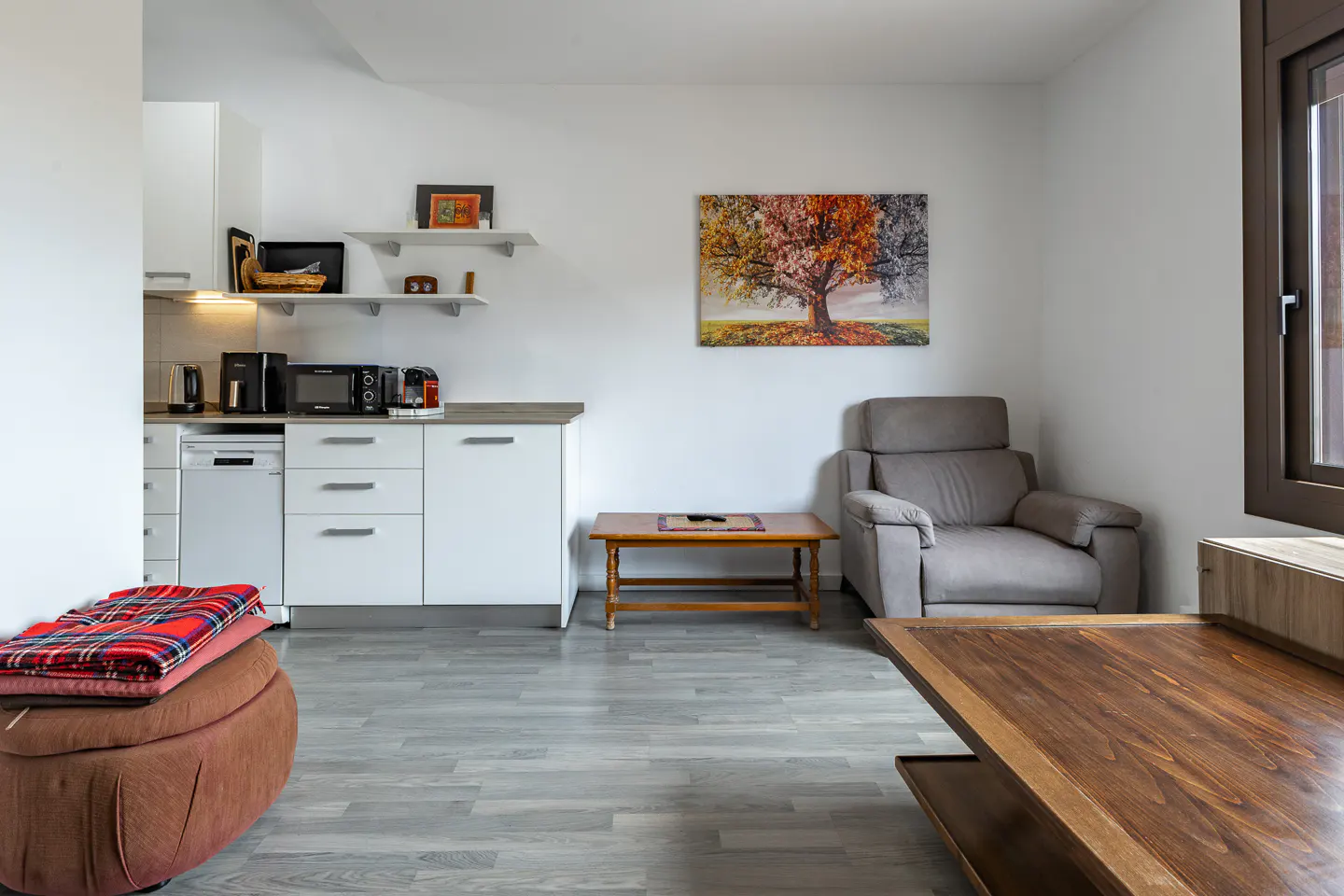 A modern apartment interior with white cabinets, a gray armchair, and a wooden table. A colorful tree painting hangs on the white wall.