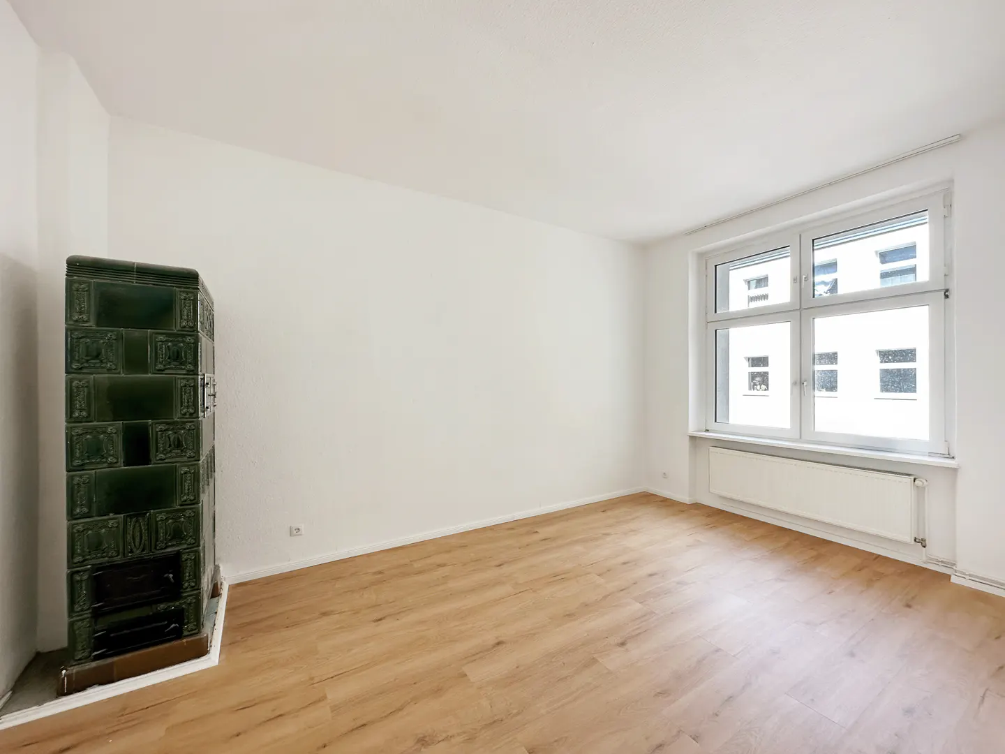 Empty room with white walls, light wood flooring, a large window, a white radiator, and a vintage green tiled stove.