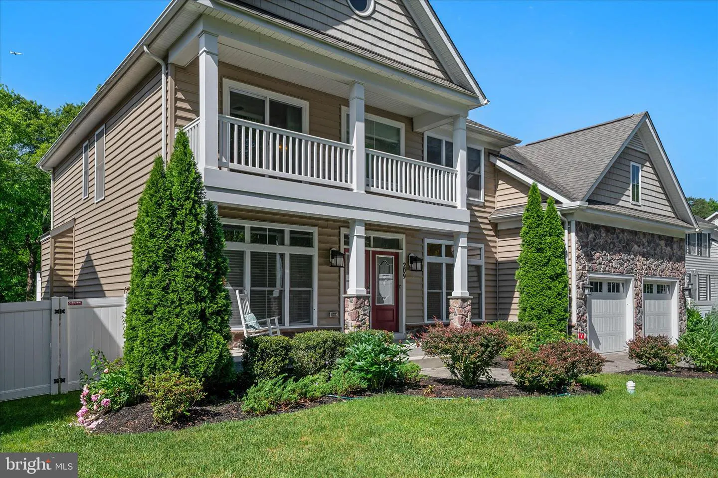 Two-story house with tan siding, white trim, and a red front door. A balcony with white railings is above the porch. Green lawn and bushes in front.