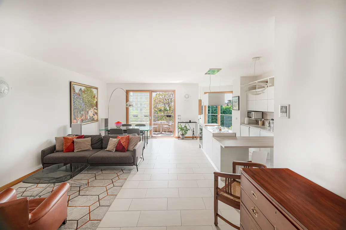 Bright, open-concept living space with a gray sofa, glass table, and white kitchen. A balcony is visible through the sliding glass doors.