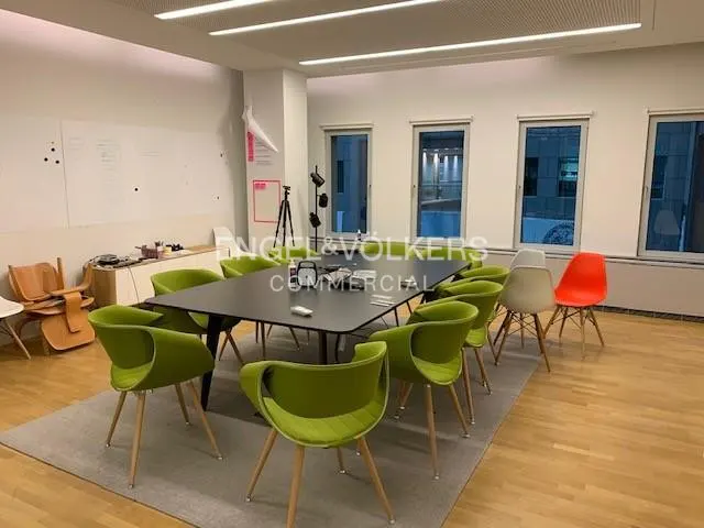 Bright office space with a black conference table surrounded by green chairs on a gray rug. Windows line the back wall.