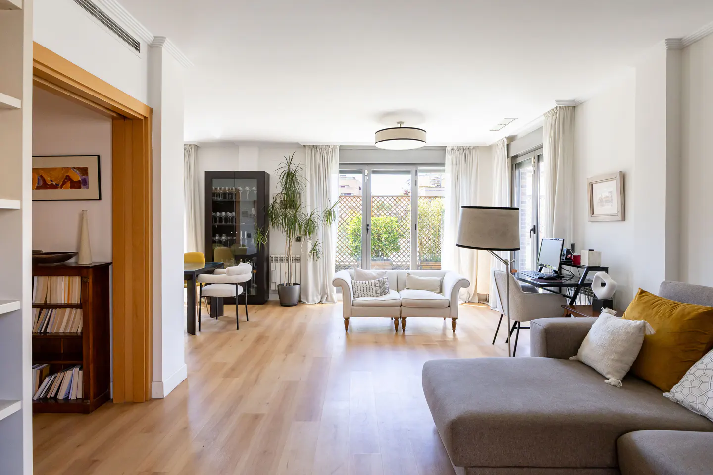 Bright, open-plan living room with hardwood floors, white walls, and large windows. A gray sectional sofa and a white loveseat provide seating.