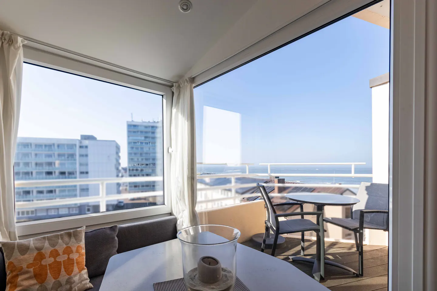Bright sunroom with a white table, gray cushions, and a view of the ocean and city buildings through large windows and an open door to a balcony.